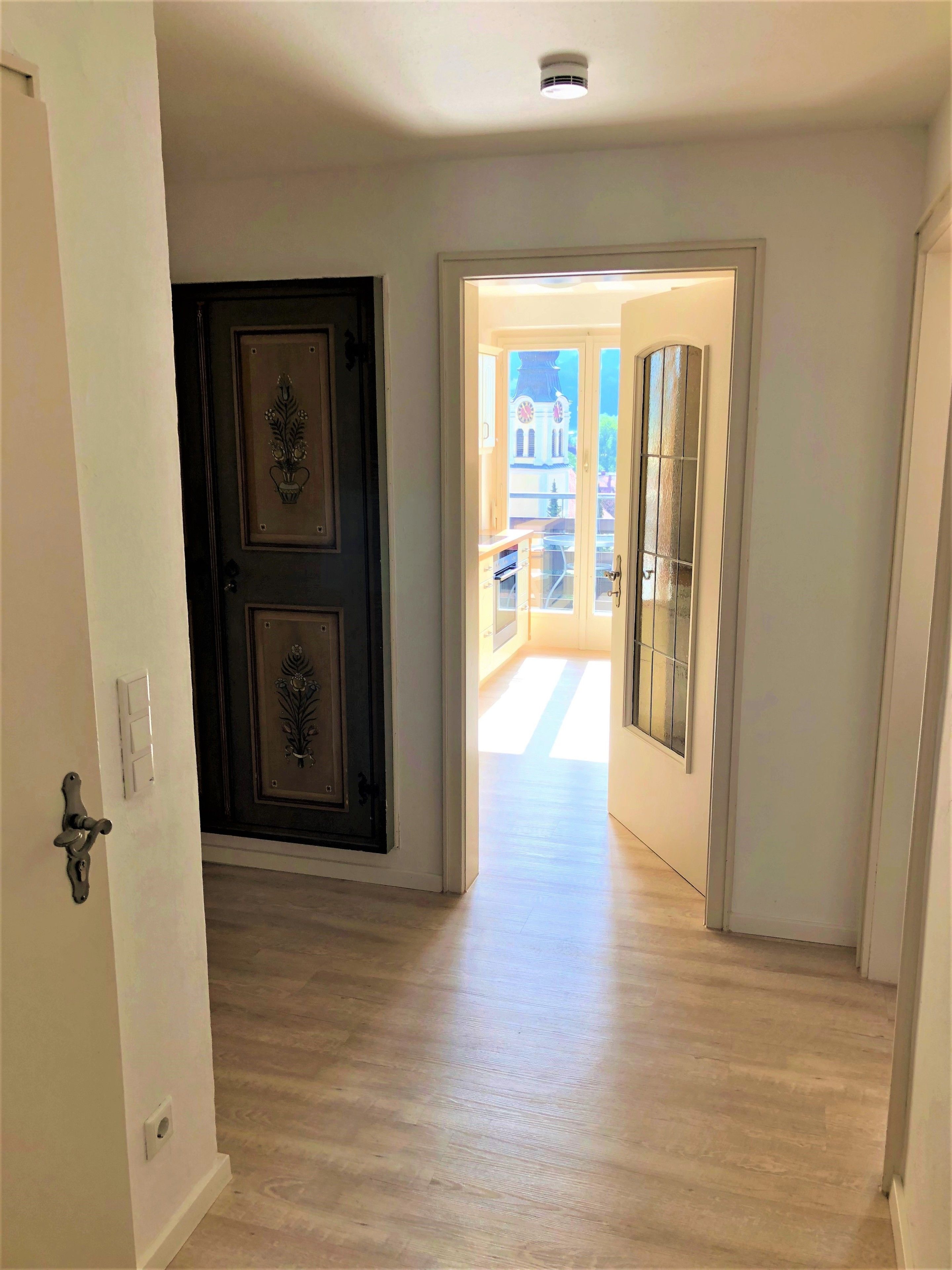 Un pasillo con piso de madera, una puerta oscura con detalles decorativos, y un vano luminoso que da a un espacio exterior con vistas a un edificio y un balcón.