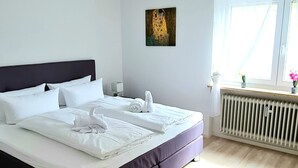 Cama bem arrumada com lençóis e toalhas brancas, cabeceira escura, radiador e janela com cortinas leves.