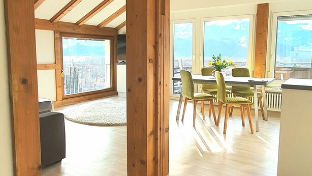 Um quarto espaçoso com vigas de madeira, área de jantar com cadeiras verdes e grandes janelas com vista.