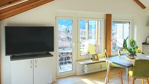 A spacious living room with a large TV, a dining area with a table and chairs, and a view of the outside through large windows. - Sanosueno 1 - Traumhafter Bergblick (Sonthofen)