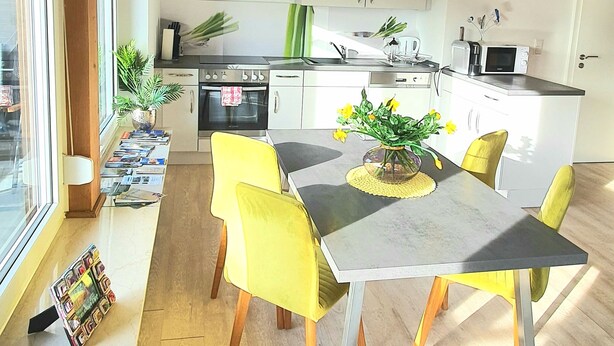 A modern kitchen with wooden beams, a dining area with yellow chairs, and a view through large windows. - Sanosueno 1 - Traumhafter Bergblick (Sonthofen)