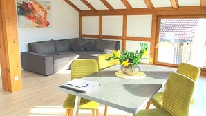 A spacious room with a high ceiling and exposed wooden beams, a dining table with chairs, a sofa, and a large window. - Sanosueno 1 - Traumhafter Bergblick (Sonthofen)