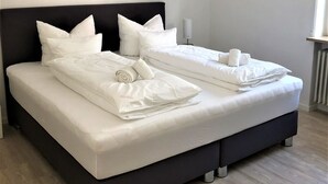 A modern bedroom with a black headboard, two white beds with neatly arranged pillows and rolled towels, a bedside table with a lamp, and a radiator on the wall. - Sanosueno 2 - Toller Bergblick (Sonthofen)