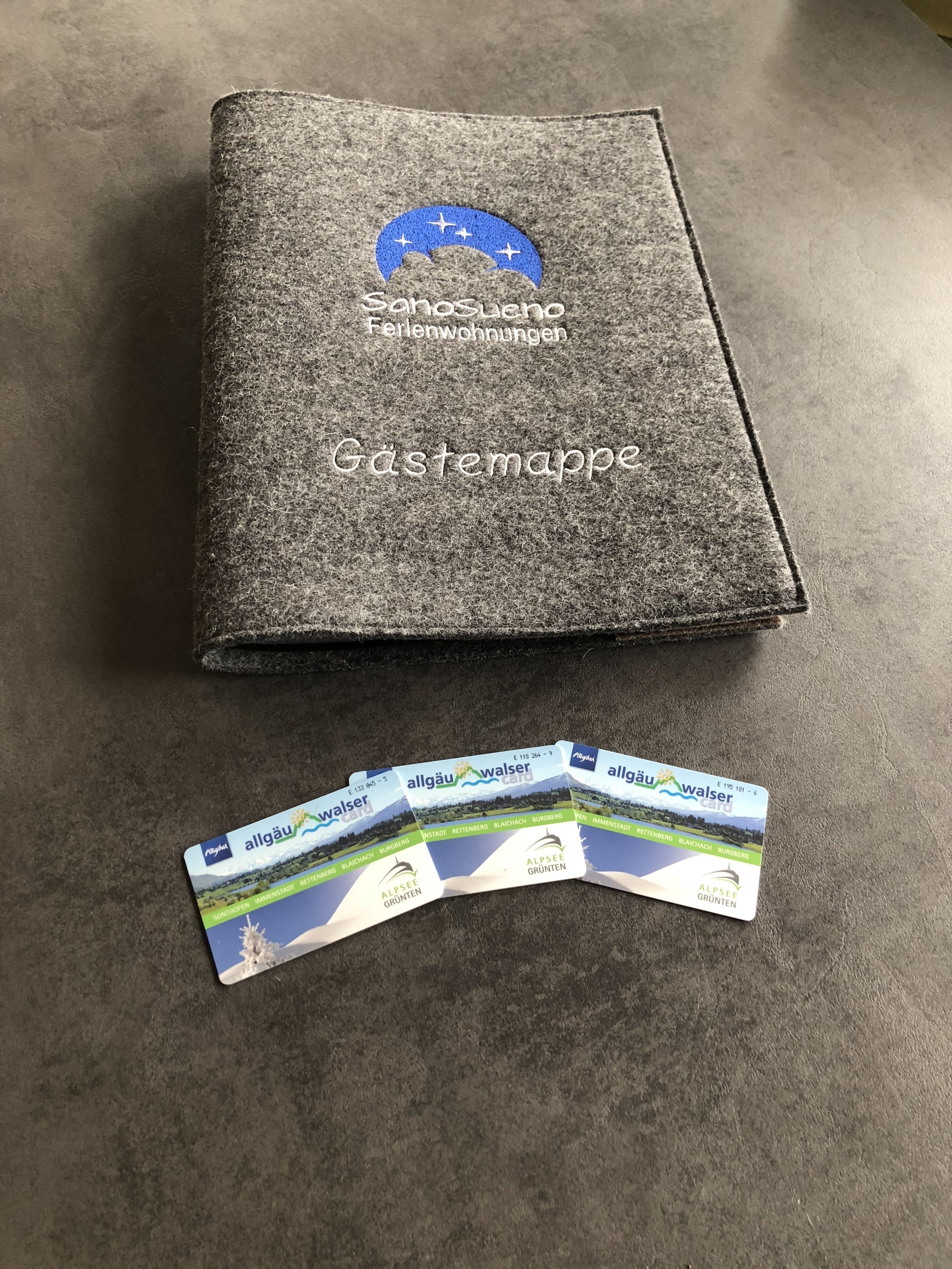 Een grijze, vilten maphoes met de tekst 'Sanosueno Ferlenwohnungen' en 'Gästermappe'. Daaronder liggen drie gevouwen kaartjes met 'allgäu walser card' en 'ALPSEE GRÜNTEN' tekst erop.