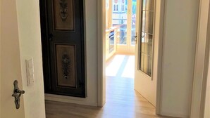 A hallway with a wooden floor, a dark door with decorative elements, and a bright doorway leading to an outdoor area with a view of a building and a balcony. - Sanosueno 2 - Toller Bergblick (Sonthofen)