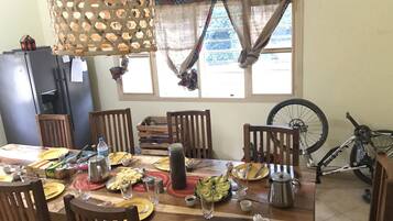 A dining area with a wooden table set for a meal, chairs, a hanging light fixture, a bicycle, and a window with curtains.