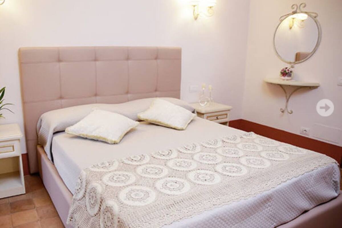 A neatly made bed with a white duvet and pillows, a bedside table with a lamp, and a wall-mounted mirror.