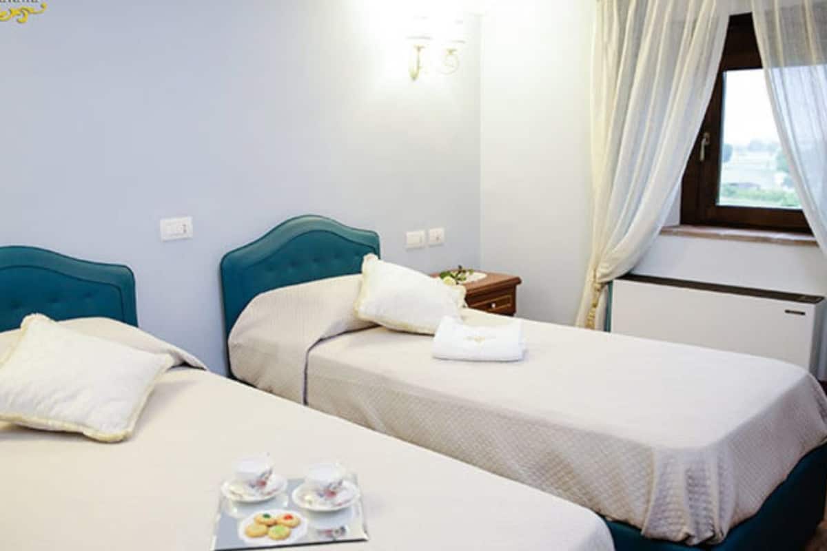 A hotel room with two beds, a small table with a tray of cookies, a nightstand, a window with curtains, and a wall-mounted light fixture.