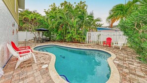 A swimming pool with a curved edge, surrounded by a brick patio. There are two red and two white lounge chairs. - Dreamscape: North End Pool Home, 2 Blocks to Beach by Anna Maria Life Vacation Rentals (Anna Maria)