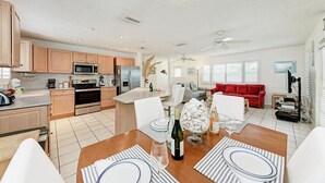 A modern kitchen with wooden cabinets, a dining area with striped placemats, and a living room with a red sofa and a ceiling fan. - Dreamscape: North End Pool Home, 2 Blocks to Beach by Anna Maria Life Vacation Rentals (Anna Maria)