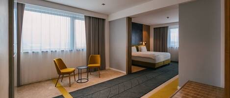 Habitación de hotel con cama, dos sillas, una mesita y un ventanal con cortinas.