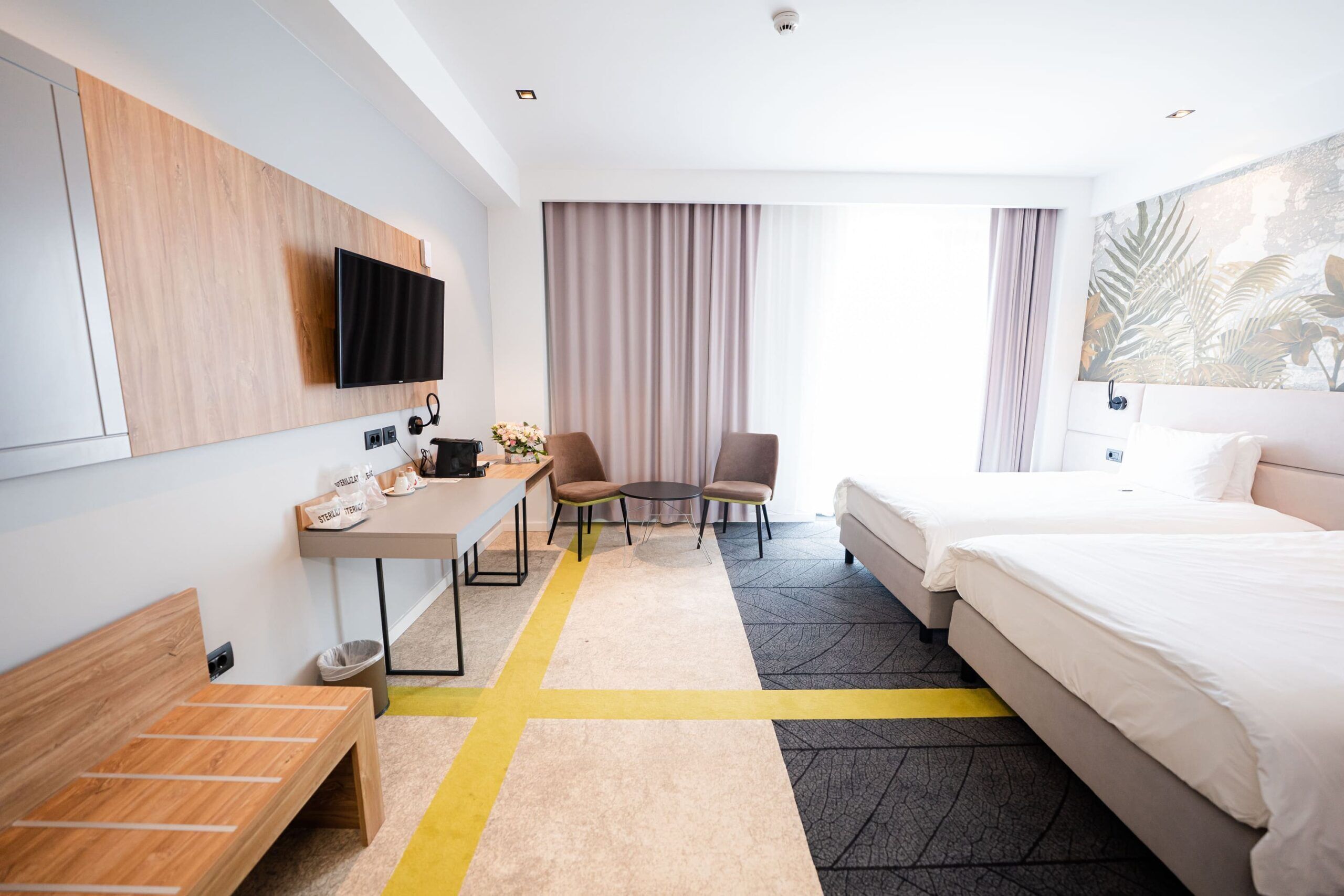 A hotel room with a bed, desk, chairs, a TV, and a large window with curtains.