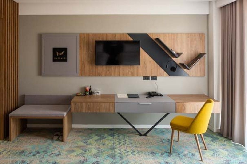 A modern hotel room with a desk, a bench, a television mounted on a wooden panel, and a colorful abstract-patterned carpet.