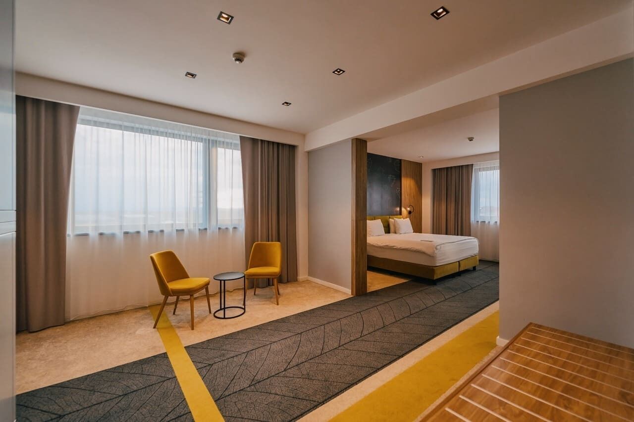 Habitación de hotel con cama, dos sillones, una mesita y vista a la ciudad a través de amplias ventanas.