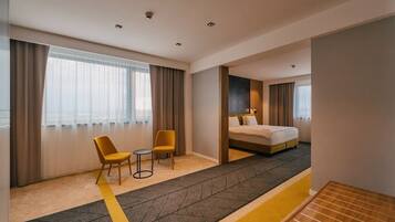 Habitación de hotel con cama, dos sillones, una mesita y vista a la ciudad a través de amplias ventanas.