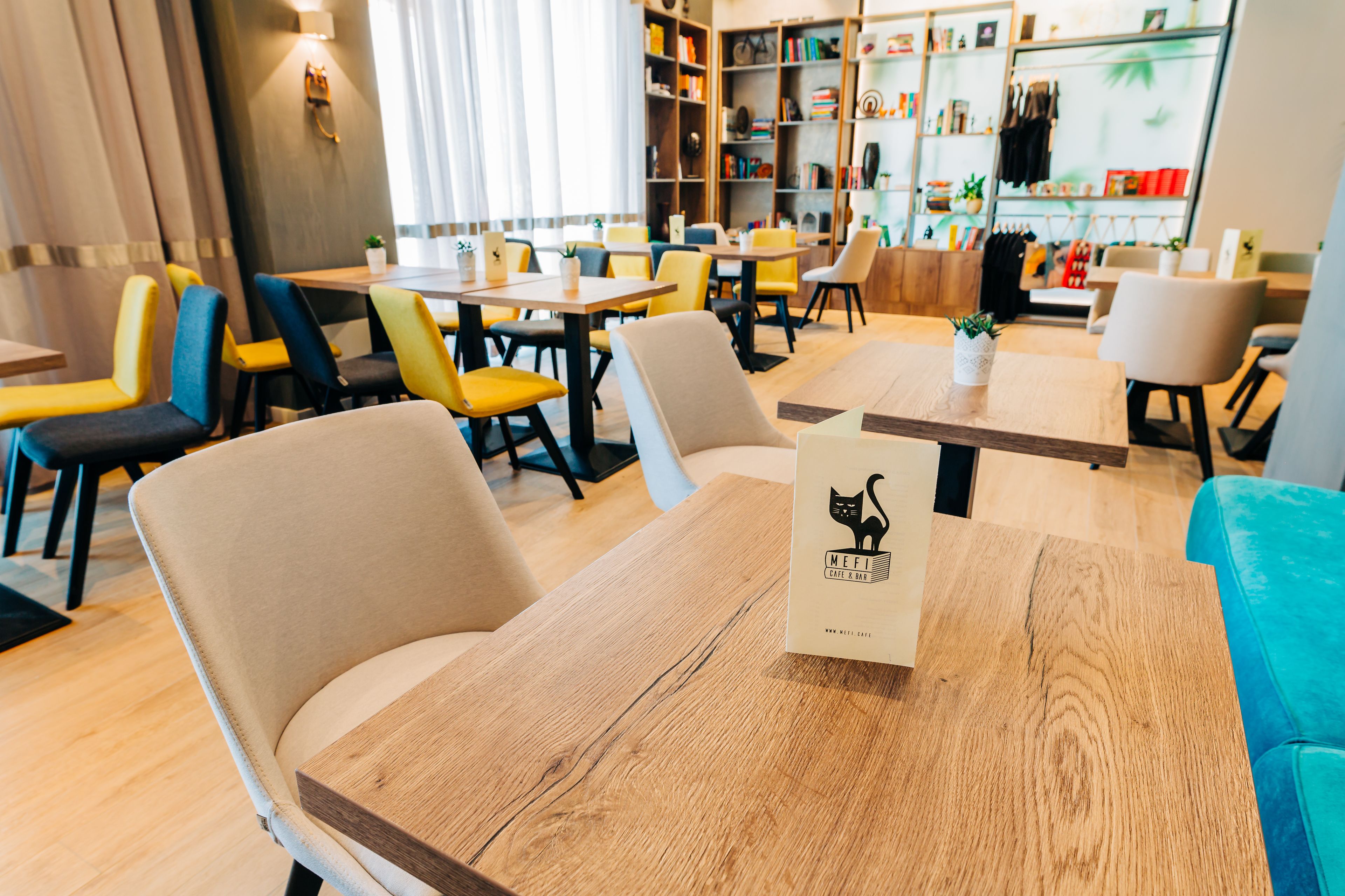 A modern cafe with wooden tables and chairs, a bookshelf with books and decorative items, and a blue sofa in the background.