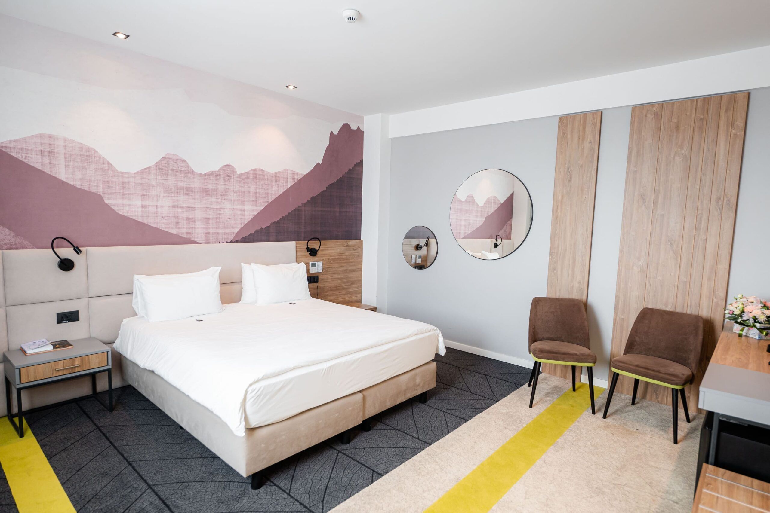 Ein Hotelzimmer mit Bett, Nachttischen, Schreibtisch, Stühlen, Spiegel und Wandgemälde.