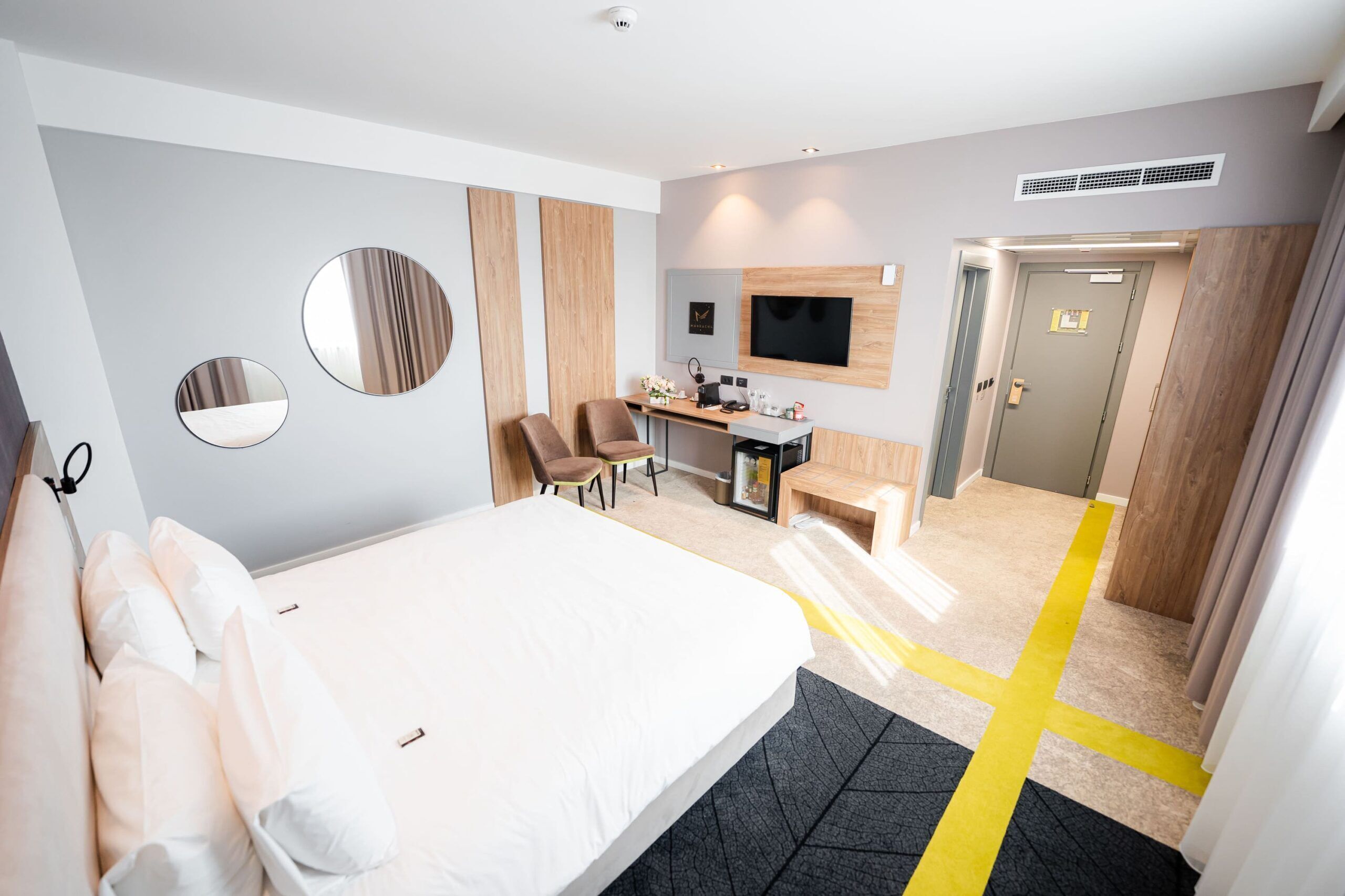 A hotel room with a bed, two chairs, a desk, a TV, and a door.