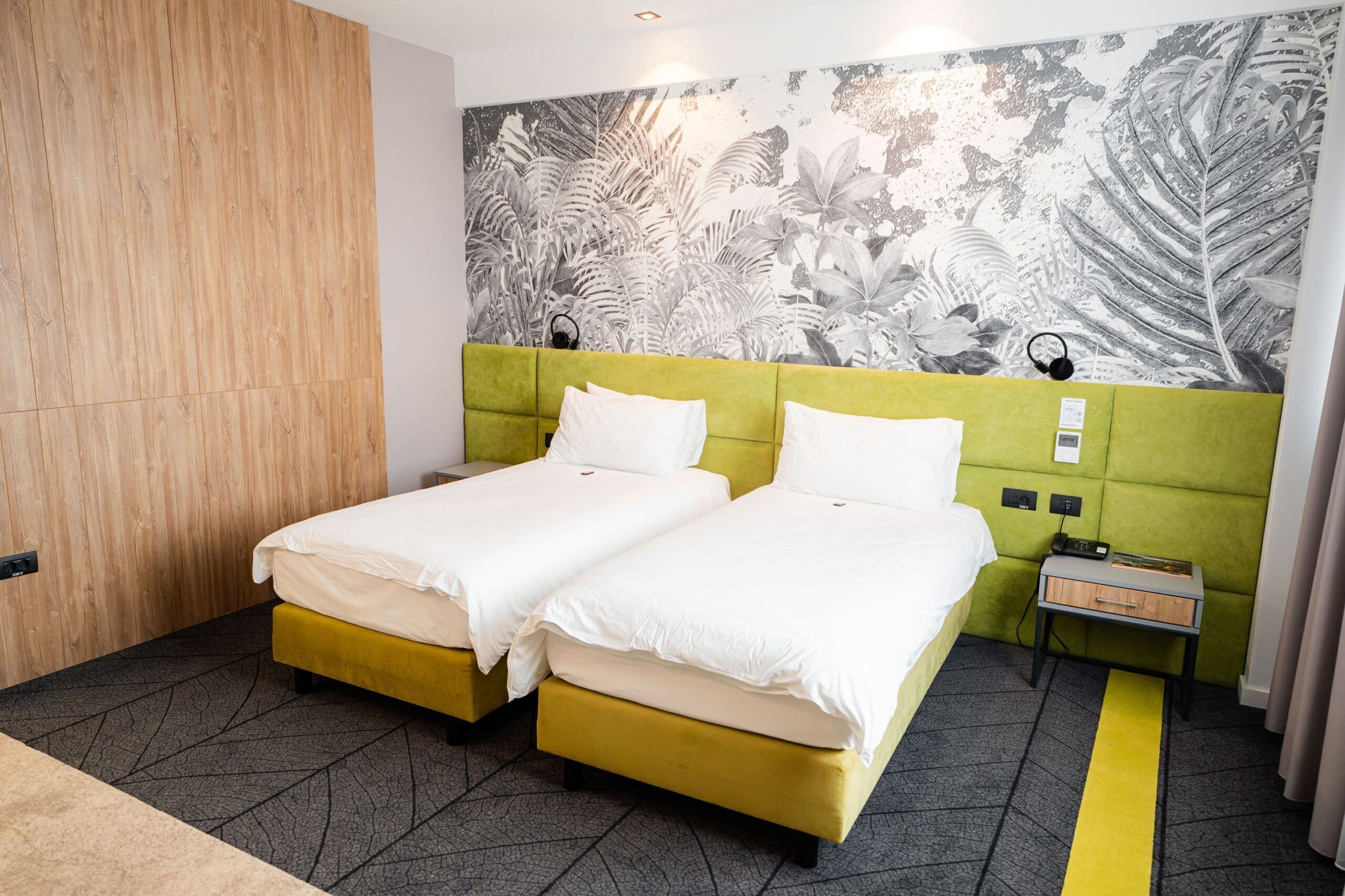 A hotel room with two beds, a green headboard, a wall with a leaf pattern, a nightstand, and a power outlet.