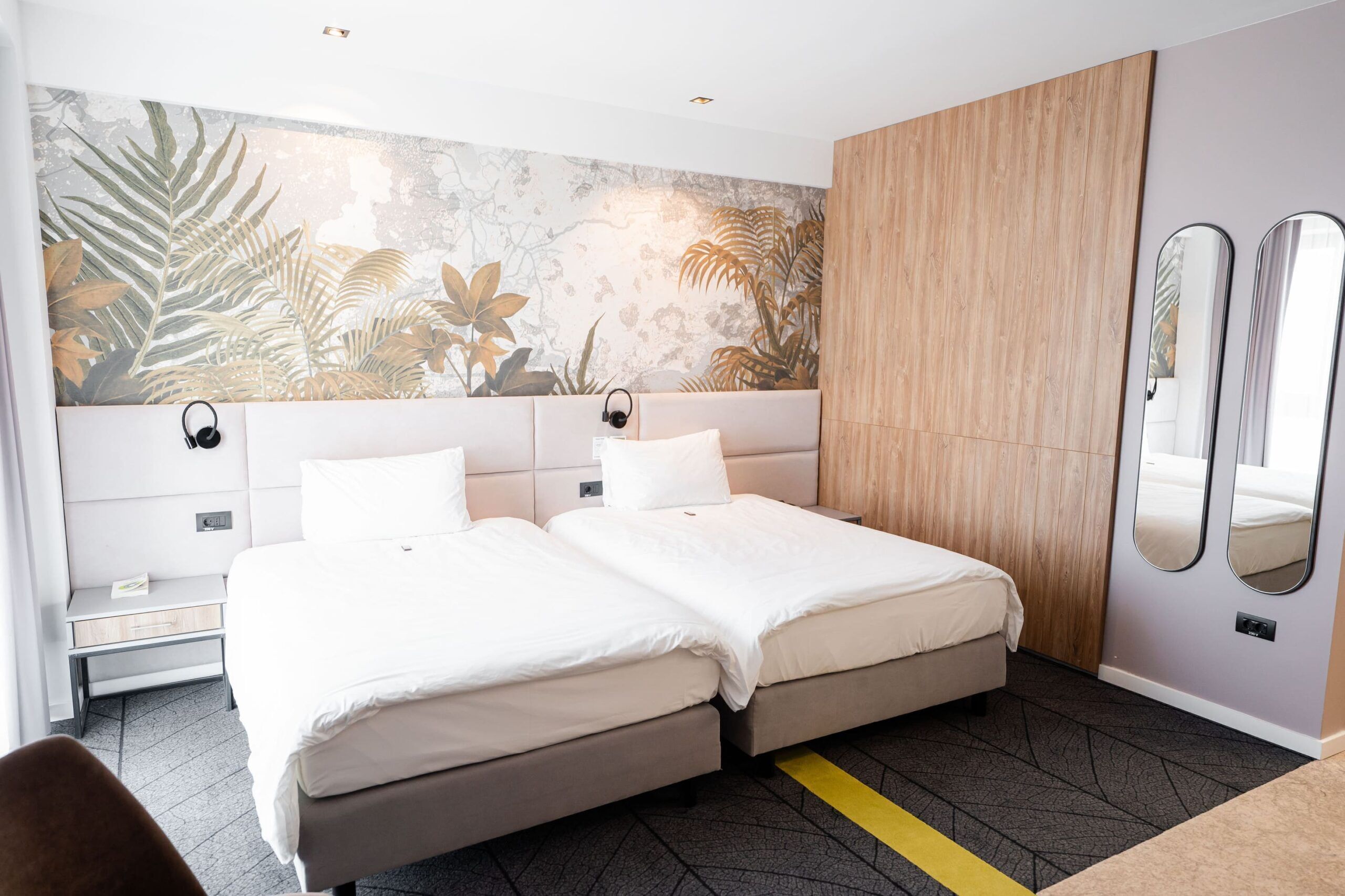 A hotel room with a bed, bedside tables, a mirror, and a wall with a tropical leaf mural.