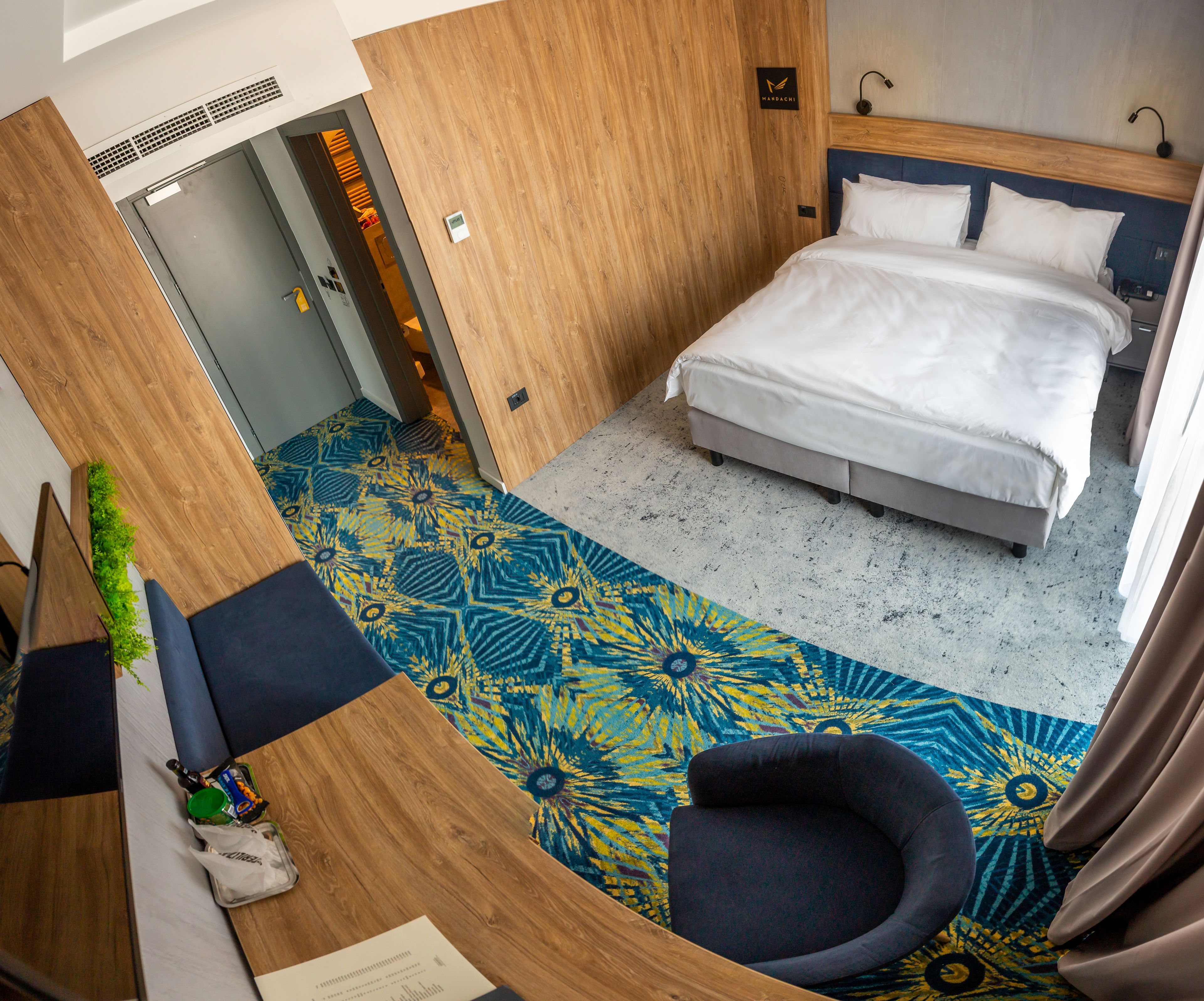 A hotel room with a bed, a desk with a chair, a patterned carpet, and a wooden wall.