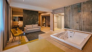 A modern hotel room with a large bathtub, a sofa, a coffee table, and a painting on the wall.
