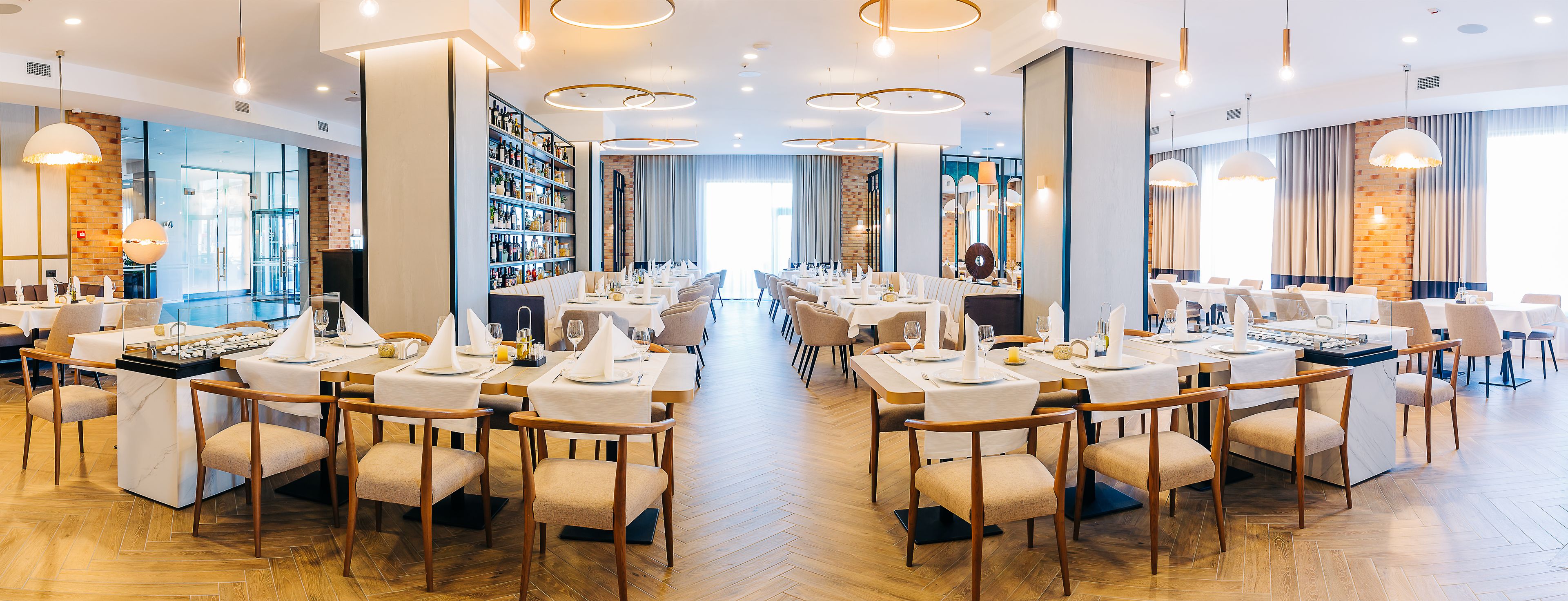 Amplia sala de comedor de hotel con varias mesas dispuestas para comer, una zona de bar con una gran selección de botellas, y vistas a la ciudad a través de grandes ventanales.