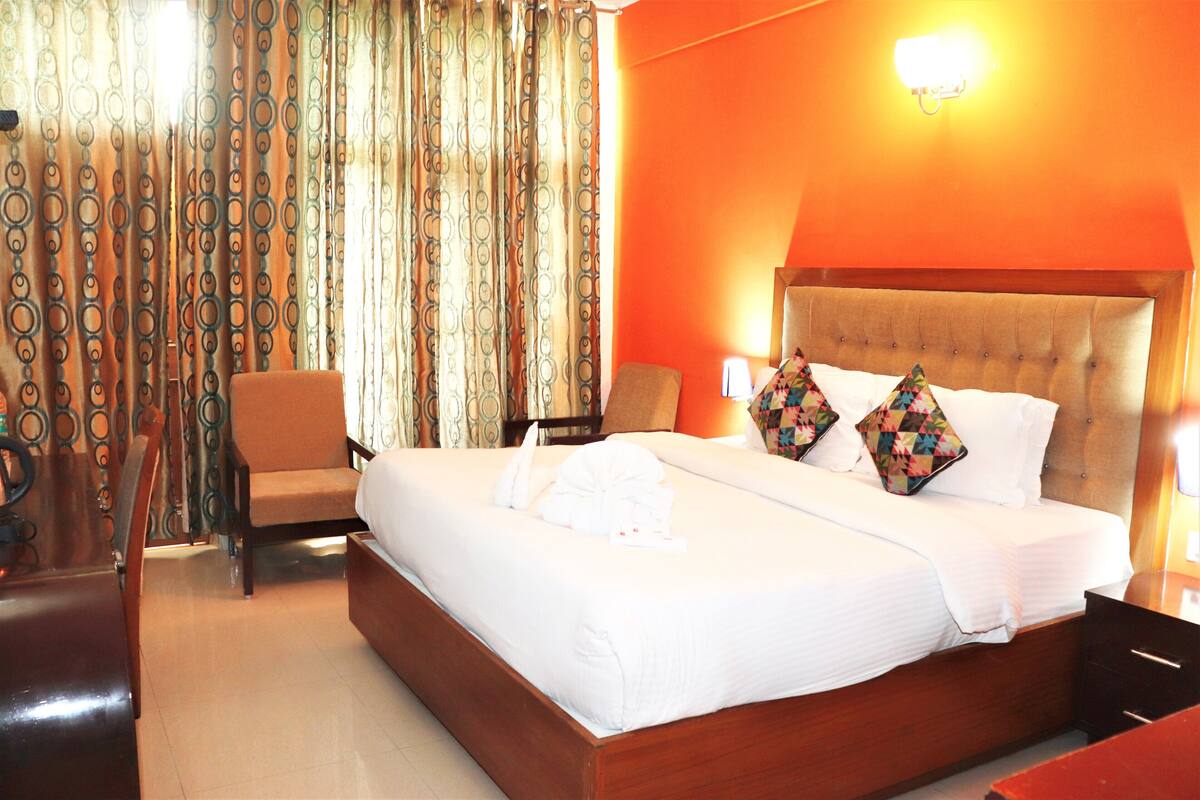 A hotel room with a large bed, a wooden headboard, two pillows, a chair, a desk, and a curtain with a circular pattern.
