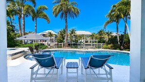 A pool area with two lounge chairs and a small table, surrounded by palm trees and a clear blue sky.