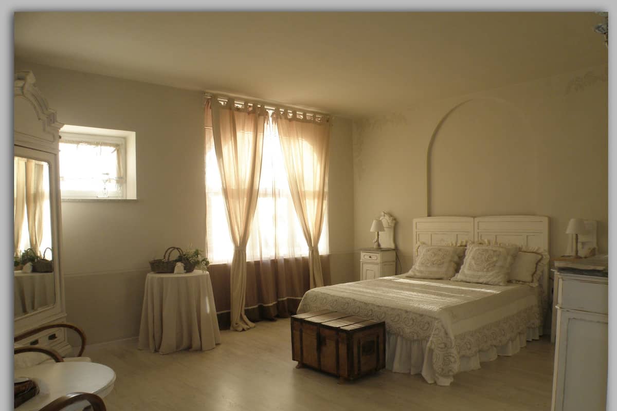 A bedroom with a large bed, a bedside table, a small table with a lamp, and a window with curtains.