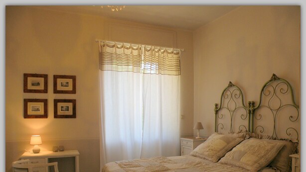 Un dormitorio con cama, una silla, una mesita de noche, una lámpara y una ventana con cortinas.