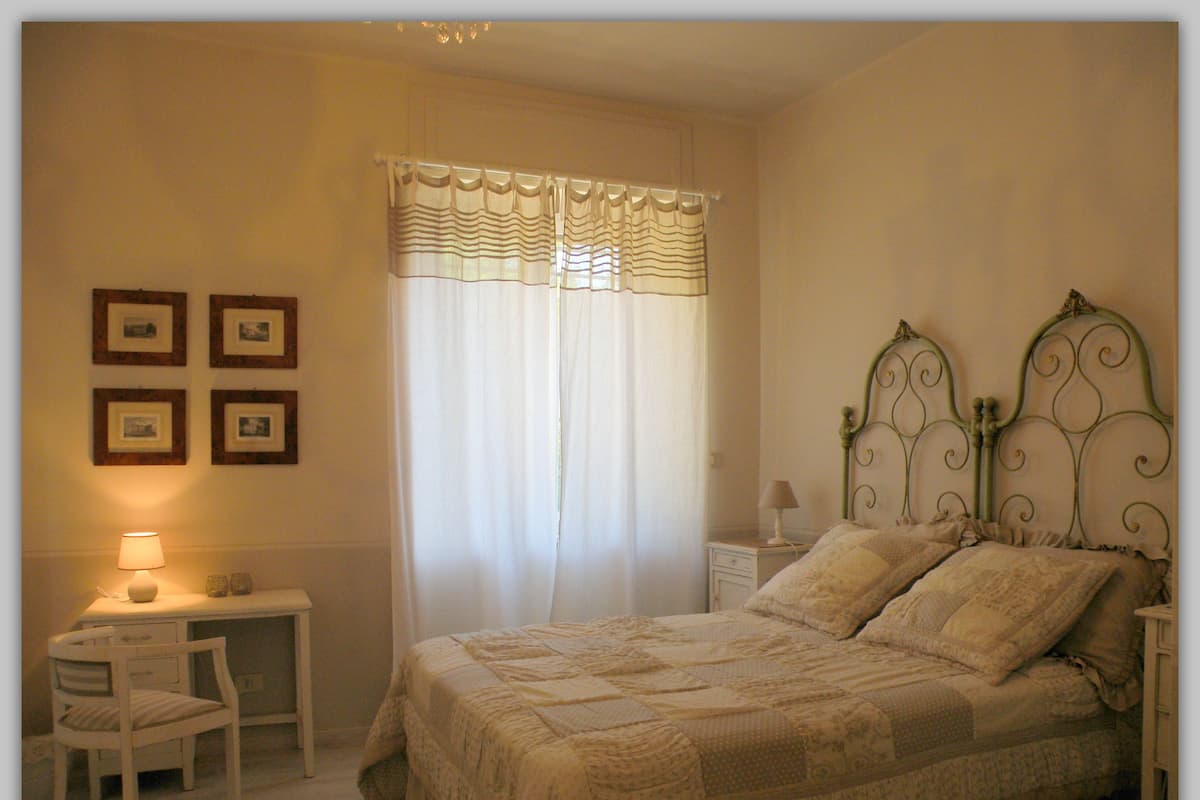 A bedroom with a bed, a chair, a nightstand, a lamp, and a window with curtains.