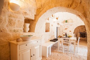 A stone-walled room with a table and chairs, a bench, and a wall-mounted shelf with decorative items.