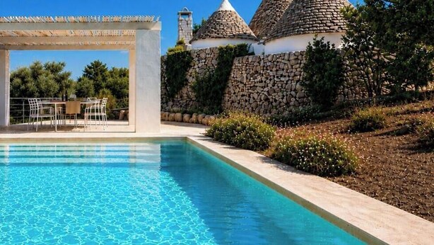 Ein Pool mit einer Pergola und Sitzgelegenheit, umgeben von Steinmauern und viel Grün.