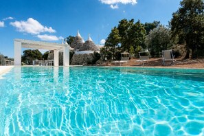 A clear swimming pool with a white pergola structure, stone buildings, and lounge chairs in the background.