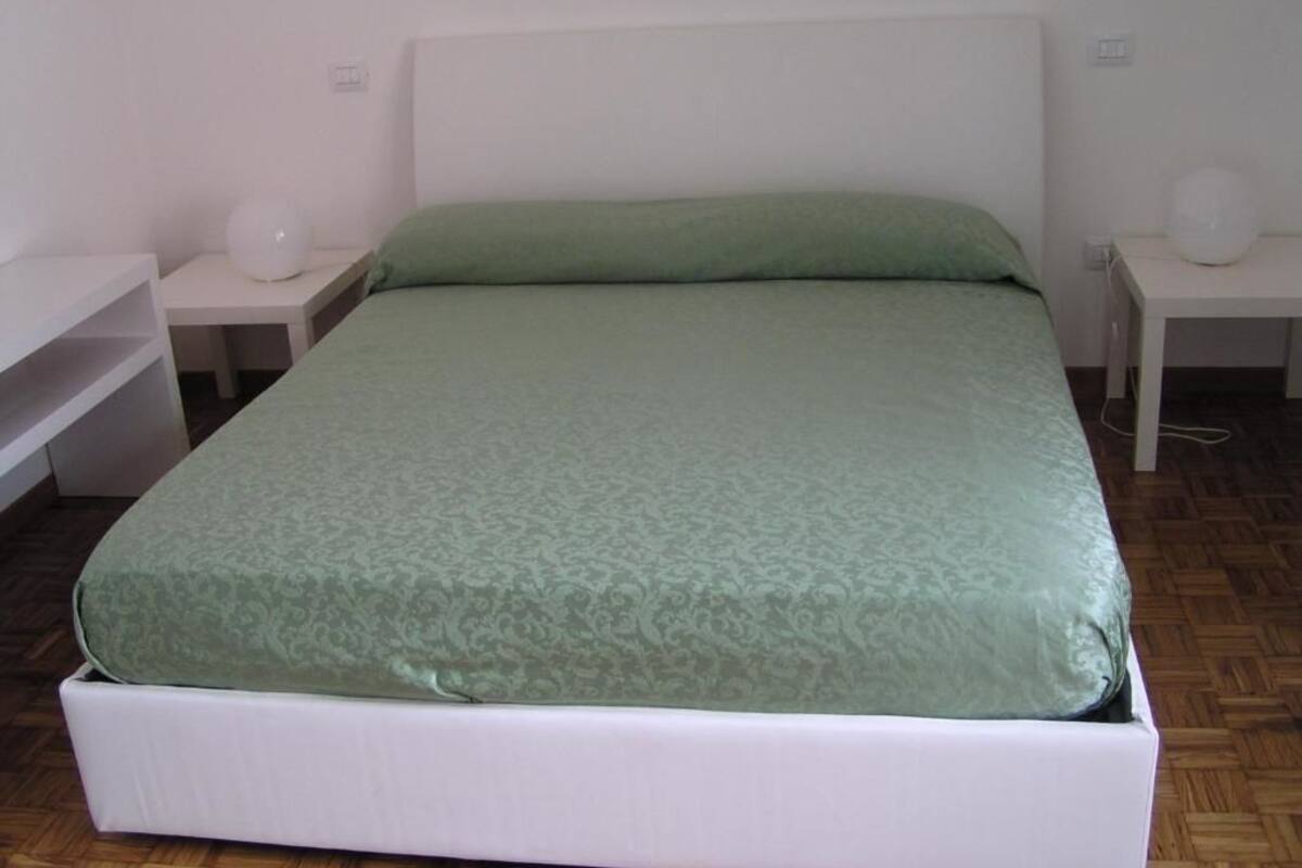 Habitación individual con una cama y cabecera blanca, una colcha verde y dos mesitas de noche con lámparas.