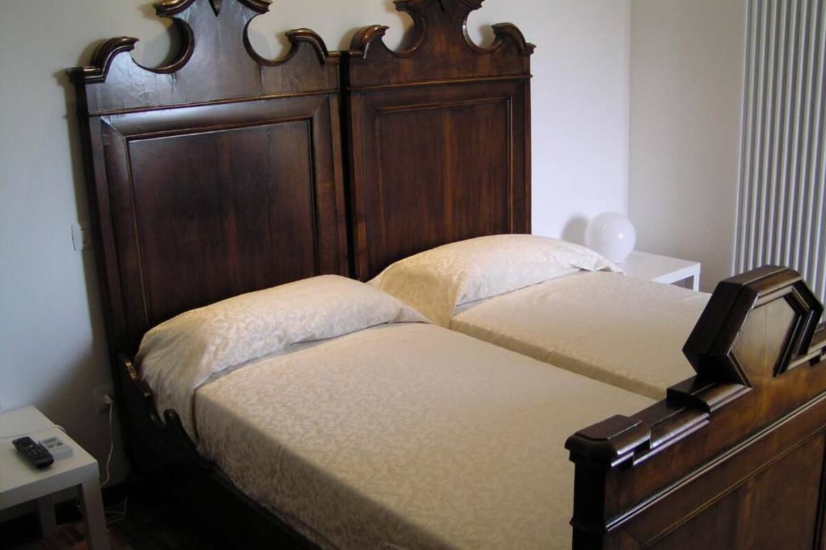 Una cama doble con cabecera de madera, una mesita de noche con lámpara y una ventana con persianas verticales.