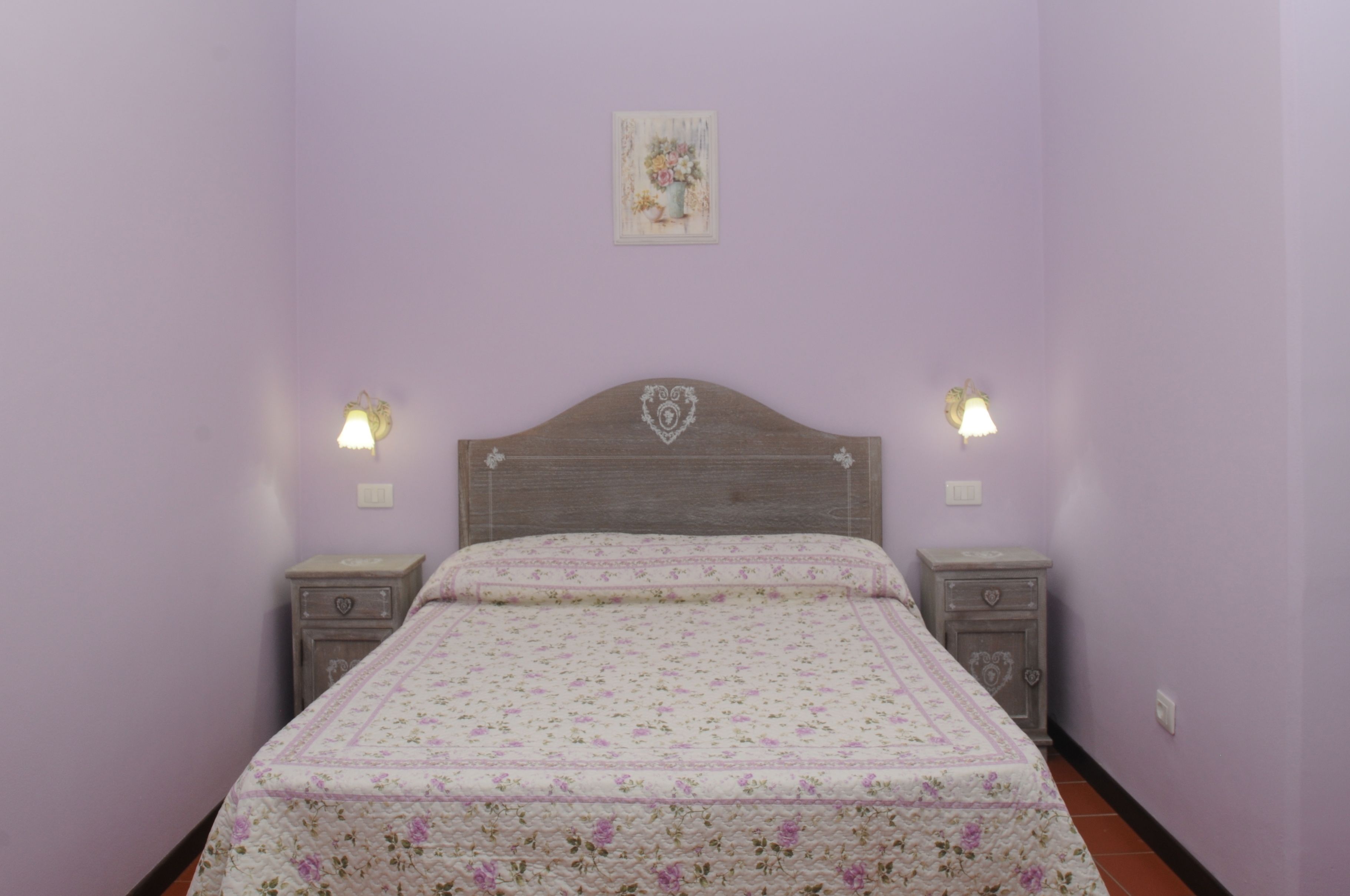Ein Schlafzimmer mit einem blumigen Bettüberwurf, einem hölzernen Kopfteil, zwei Nachttischen mit Lampen und einer kleinen Wanddekoration.