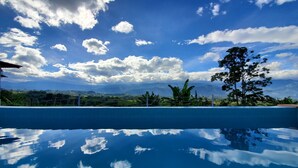 Cielo azul despejado con nubes, piscina que refleja el cielo y follaje al fondo.