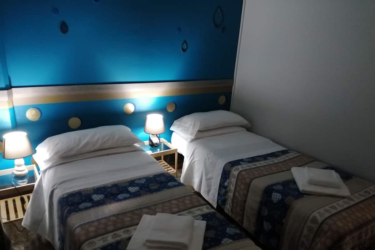 A hotel room with two single beds, each with a nightstand and a lamp. The room has a blue accent wall with circular decorations.
