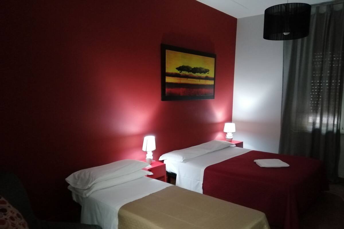 A hotel room with two beds, a red wall, a painting, and a window with curtains.