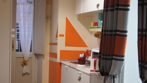 Ein schmaler Flur mit einer zur Kammer führenden Tür, einer Küchenzeile mit orangefarbenen Akzenten und einem Streifenmuster-Vorhang.