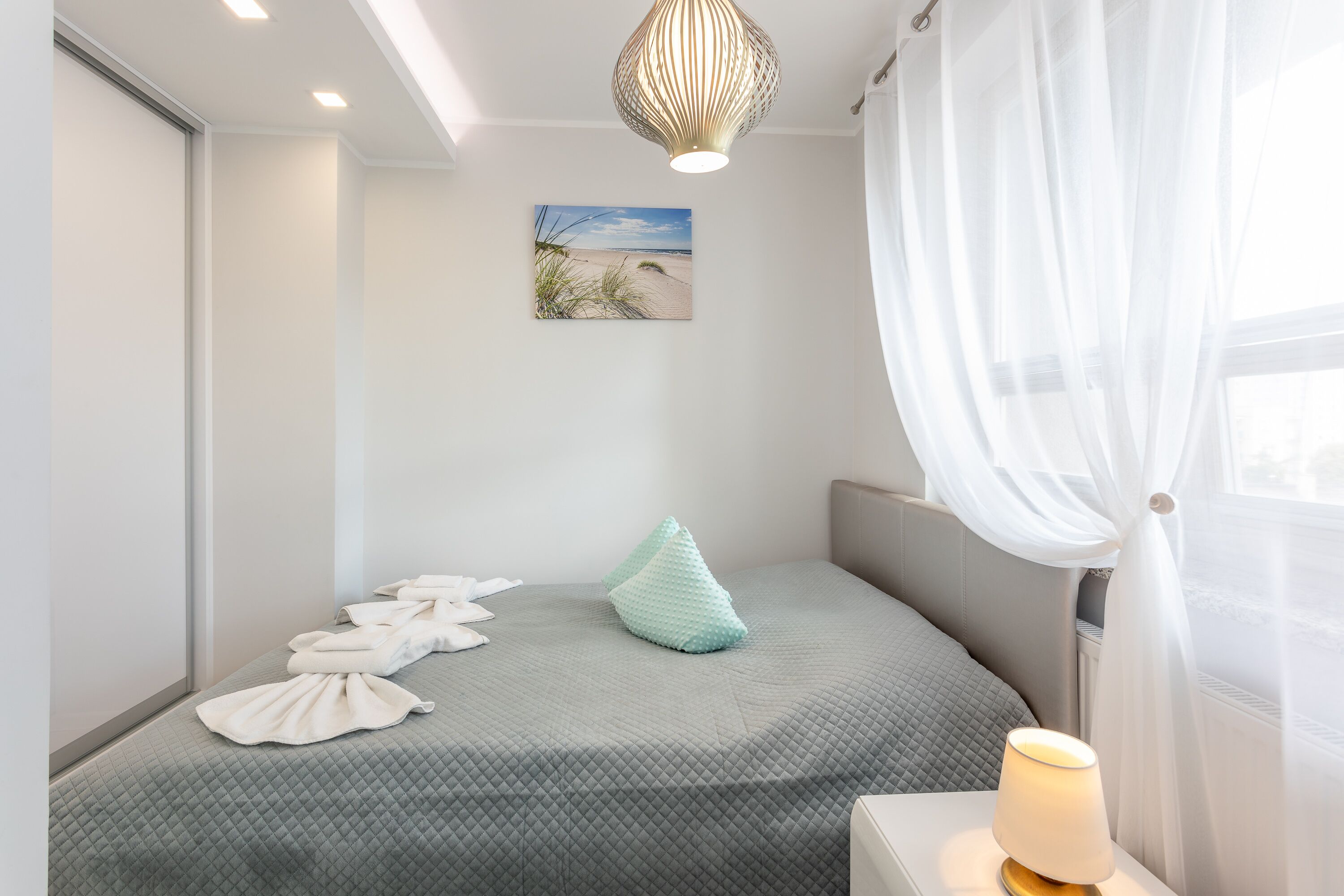 Un letto rifatto con copriletto grigio trapuntato, un cuscino azzurro e diversi asciugamani bianchi piegati. Sopra il letto, un quadro raffigura una spiaggia.
