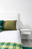 A bed with a white headboard, a green pillow, and a white pillow. There is a white nightstand with a white lamp beside the bed.
