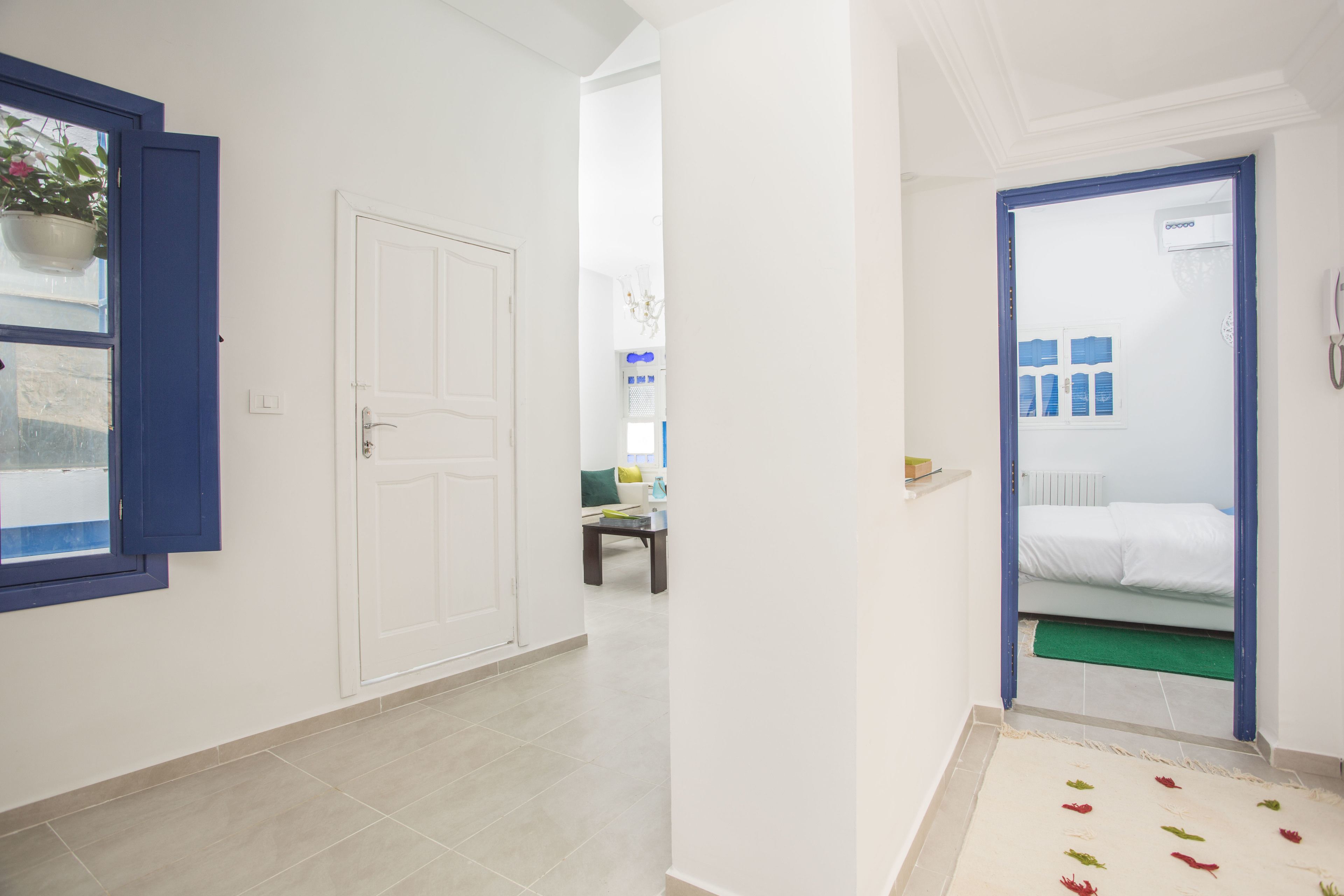 A hallway with a white door, a blue-framed window, and a bed with white bedding in the adjacent room.