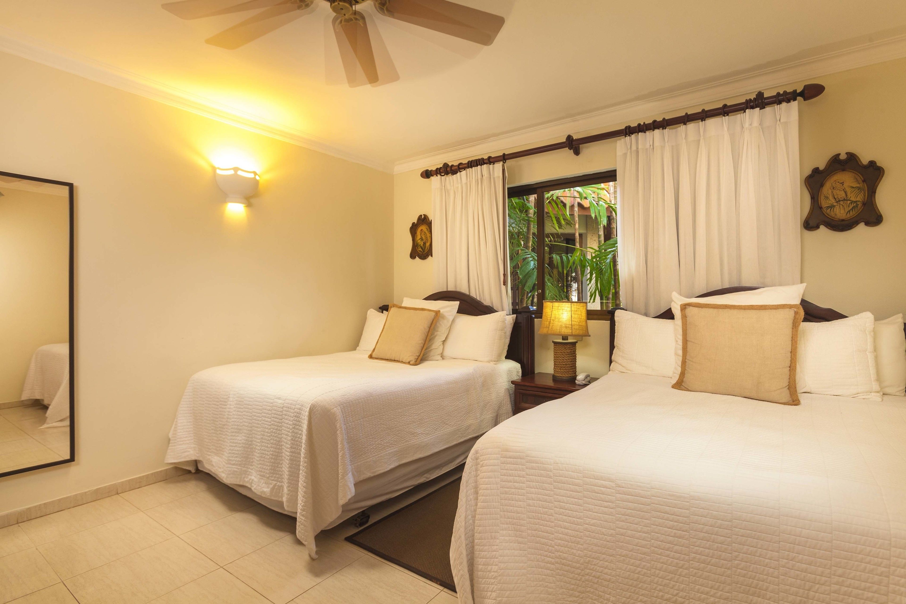 A hotel room with two beds, a ceiling fan, a window with curtains, a bedside lamp, and a wall-mounted clock.