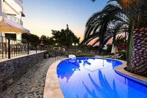 A swimming pool with a stone border, a palm tree, and a building with a balcony. - Kamelia Apartments (Mylopotamos)