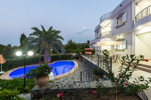 A pool area with a stone deck, palm trees, and a building with balconies and outdoor seating. - Kamelia Apartments (Mylopotamos)