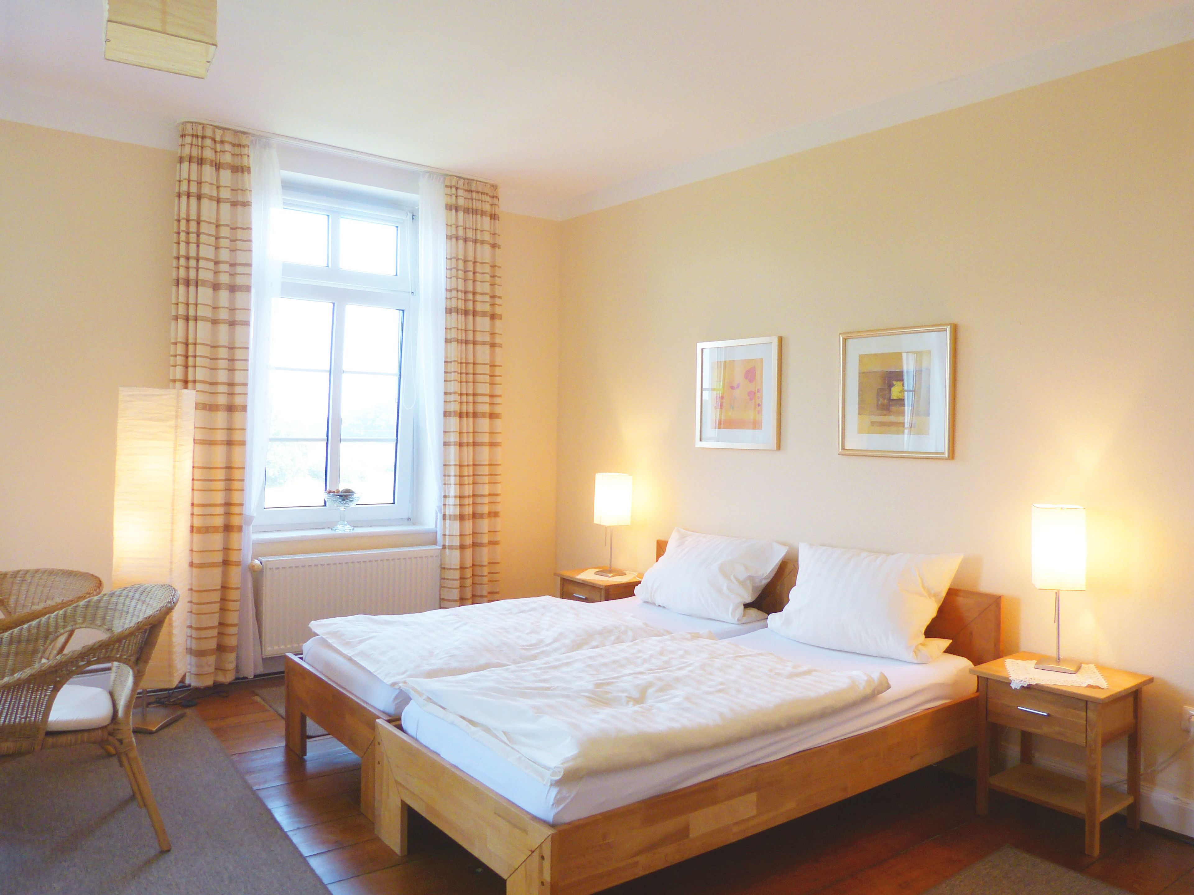A bedroom with a double bed, two bedside tables with lamps, a chair, a window with curtains, and two framed pictures on the wall.