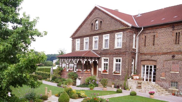 A two-story brick building with a traditional design, featuring a covered porch with flower boxes and a well-maintained garden with various plants and flowers. - Bleckmanns Hof (Werne)